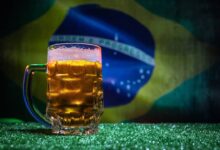 Caneca de cerveja gelada com espuma sobre grama, bandeira do Brasil desfocada no fundo.
