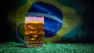 Caneca de cerveja gelada com espuma sobre grama, bandeira do Brasil desfocada no fundo.